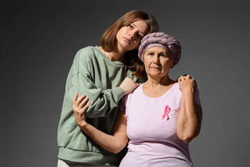 Fototapeta premium Senior woman after chemotherapy with pink ribbon and her granddaughter hugging on dark background. Breast cancer awareness concept