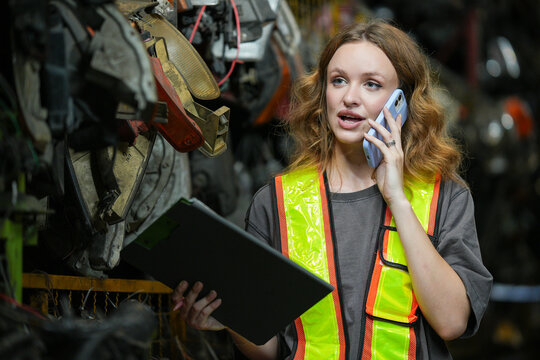 Warehouse Manager: Determined young woman, wearing safety vest and holding a clipboard, speaks on her phone amidst shelves filled with automotive parts. Scrap Yard recycle auto engine workplace