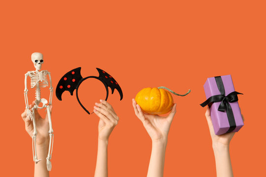 Female hands with pumpkin, skeleton, bat wings headband and gift box on orange background. Halloween celebration