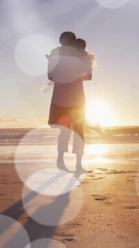 Animation of light spots over happy african american groom lifting bride on sunset beach at wedding