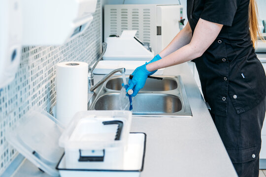 Gloved hands wash medical mirror tools under running water stream at double sink basin in clinic. Dental nurse at work, dental assistant job, healthcare worker daily routine.