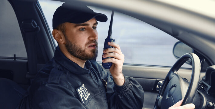 Young police officer in uniform communicating with a walkie talkie while driving a patrol car, law enforcement and public safety