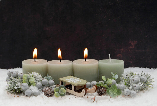 Dritter Advent. Modernes Adventsgesteck mit Eukalyptus, salbeigr&uuml;nen Kerzen,  kleinem Holzschlitten und Kunstschnee vor dunklem Hintergrund mit Textfreiraum.