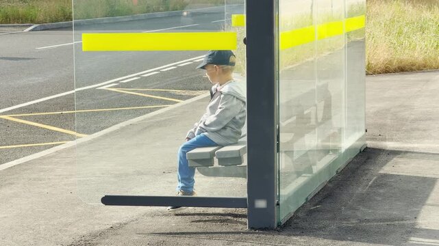 A young boy sits patiently waiting on a bench inside a glass bus stop by the road. This quiet scene feels lonely, pensive, and calm.