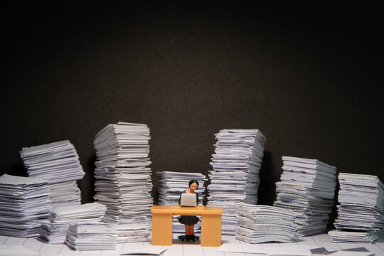 Miniature tiny people toy figure photography. Overload and work overtime concept. A female businesswomen seat on a desk with pile of document, working until night at dark room with spotlight
