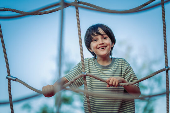 A smiling boy climbs on a rope net at the playground in summer