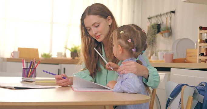 Mother guiding and helping young daughter writing homework in notebook at study desk.