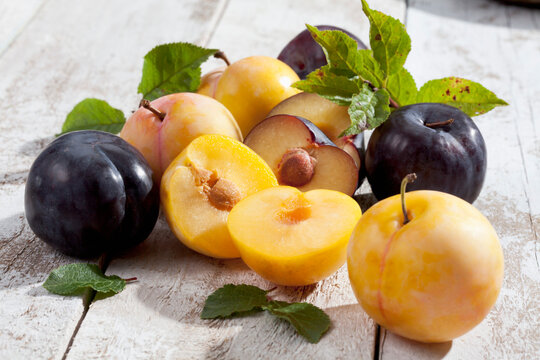Sliced and whole yellow and black gages (Prunus domestica subsp. italica) on white wooden table, studio shot