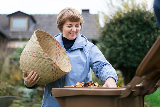 Smiling woman puts autumn leaf waste into a compost bin in the garden