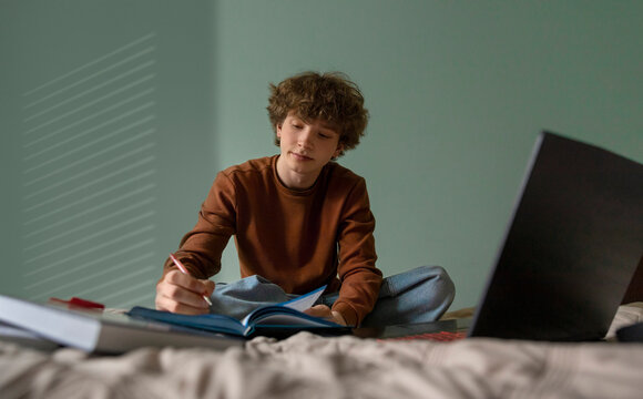 A teenage boy studies with a laptop and textbook on his bed