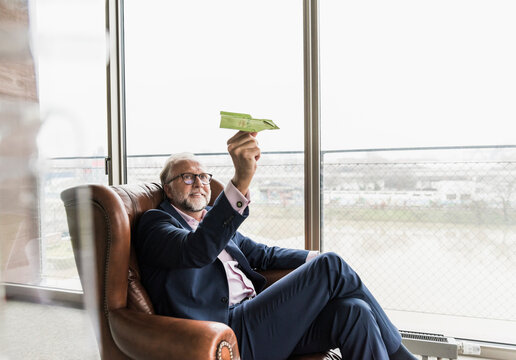Smiling mature businessman sitting in leather armchair holding paper plane