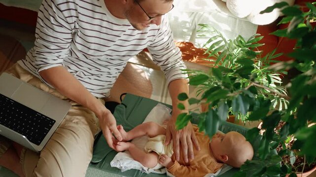 Young freelancer father combining remote work and childcare, affectionately massaging his newborn baby's feet while the infant rests in a bouncer next to his laptop in a cozy home office