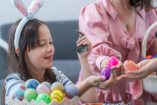 A cute mixed race girl in bunny ears and a blue striped shirt holding a bright pink painted Easter egg up beside her cheek against a bright white wall in a light-filled room at home.