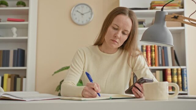 Woman writing in notebook while holding smartphone at desk in building with bookshelves, lamp and clock visible; concentration study.