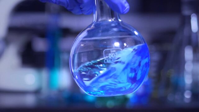 A scientist in blue gloves holds a round laboratory flask containing vibrant blue liquid,.