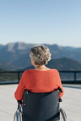 Fototapeta premium Rear view of an elderly woman in a wheelchair facing mountain vista on a balcony for serene background and healthcare design use