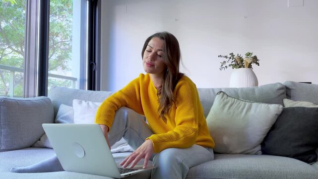 Young woman working on her laptop on the couch, then closes it to relax and thoughtfully look out the window.