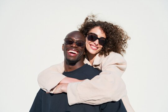 Smiling mixed couple piggyback embrace wearing sunglasses for lifestyle banner and social media background