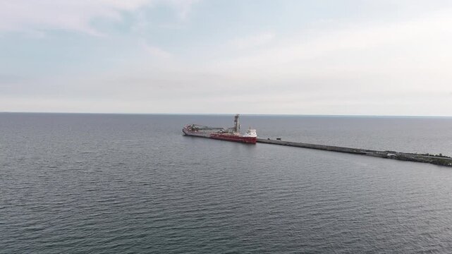 Wide aerial tracking shot moving forward over Lake Ontario toward the red hull and conveyor boom of the CSL Tadoussac bulk carrier docked at the Ash Grove cement plant pier in Mississauga, Canada