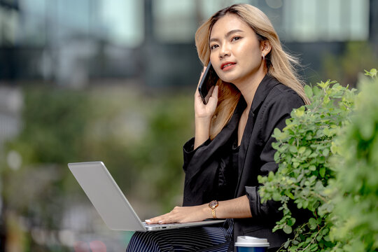 Close up of an Asian businesswoman with long blonde-dyed wavy hair speaking animatedly on her phone with mouth wide open, a laptop softly blurred in front of her outdoors.