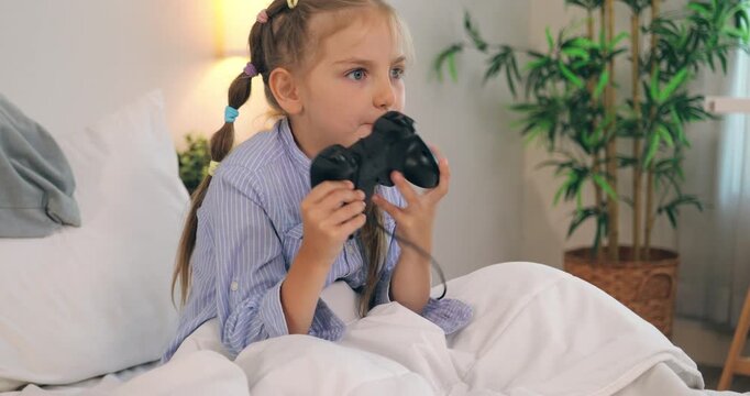 A cute little girl sitting in bed under a blanket and excitedly playing video games with a controller.