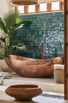Hand-hammered copper freestanding tub beside terracotta vessel on limestone ledge, featuring emerald zellige wall and tropical foliage in contemporary bathroom