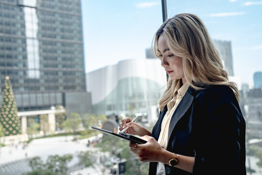 Asian businesswoman studies her tablet with a stylus poised, standing beside a glass wall as a landscaped plaza and cluster of skyscrapers stretch out in the background.