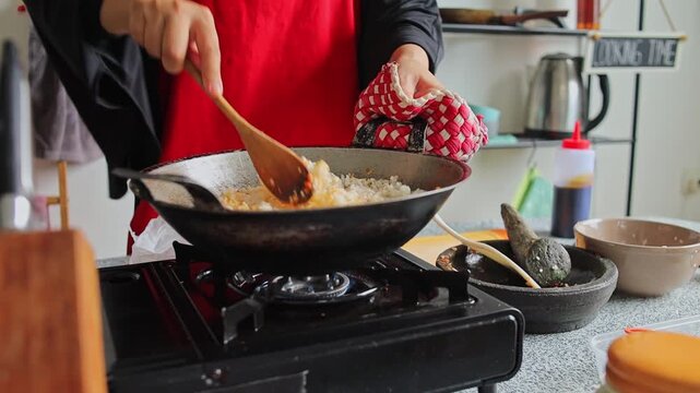 Cook holds hot wok with oven mitt while stirring rice and chili spices together using wooden spatula