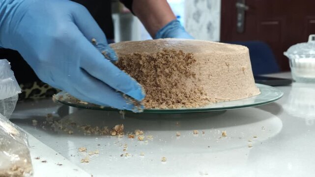 Close view shows gloved hand pressing crumb topping onto beige frosted cake on glass plate inside cozy kitchen, with gentle motion, neutral tones, and detailed dessert preparation, with cake