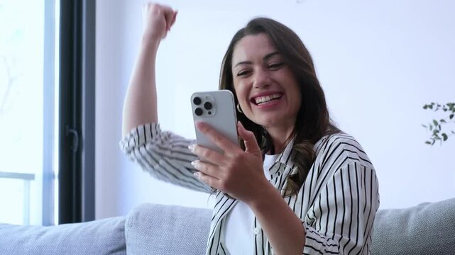 A happy young woman celebrating online success with her smartphone at home, cheering with excitement after receiving good news or winning something, portraying joy and achievement.