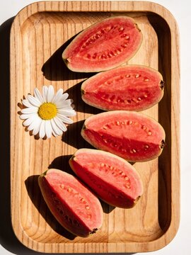 A vibrant and sun-drenched flat lay featuring freshly sliced Psidium guajava (pink guava) arranged elegantly on a rustic wooden tray. The striking contrast between the rich, ruby-pink flesh