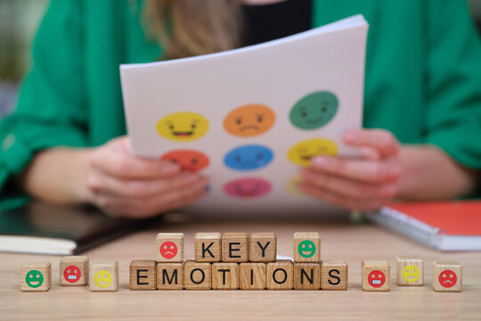A person sits at a table engaged in a creative workshop, holding a sheet with vibrant emotion faces. Wooden blocks display the words key emotions, inviting reflection on feelings.