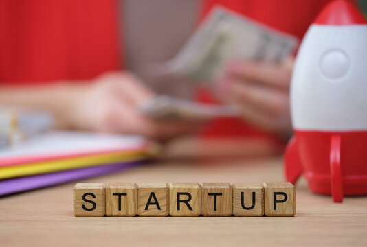 In a lively office, a person holds cash while planning for a startup, surrounded by colorful documents and a playful rocket model, symbolizing ambition and growth.