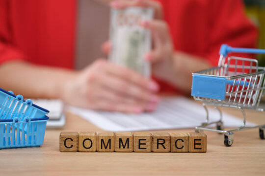 On a wooden table, playful toy shopping carts and small blue baskets surround wooden blocks spelling commerce.