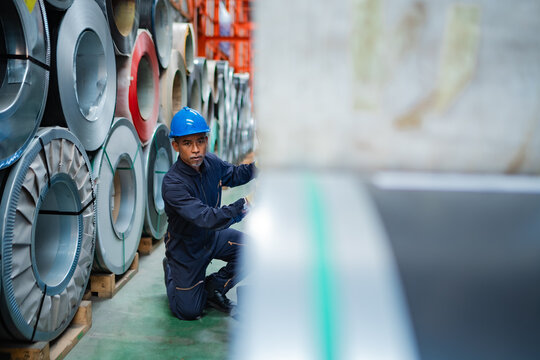 Factory worker checking stacked steel coils in warehouse aisle, inspecting material condition, inventory storage, metal products and industrial manufacturing operations.