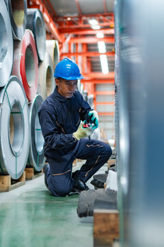 Factory worker checking steel coil inventory with handheld radio, managing warehouse communication, material inspection, production support and industrial operations.