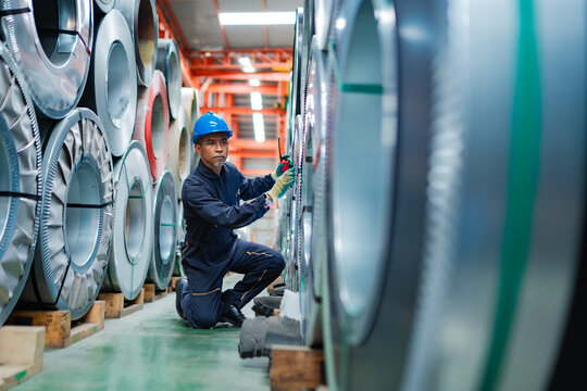 Industrial worker using handheld radio while checking steel coil inventory in factory warehouse, supporting inspection, communication, material control and production operations.