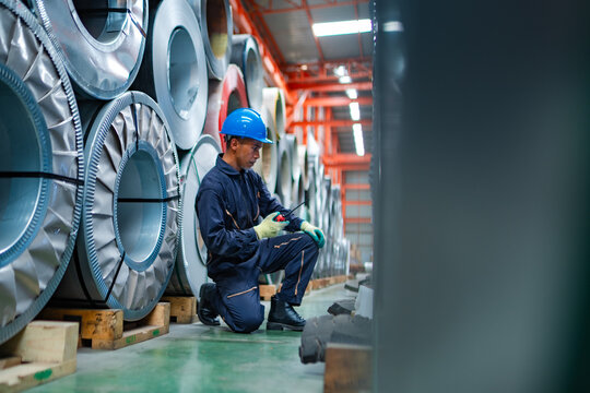 Industrial worker using radio between stacked steel coils in warehouse, checking material stock, inventory condition and coordinating factory operations.