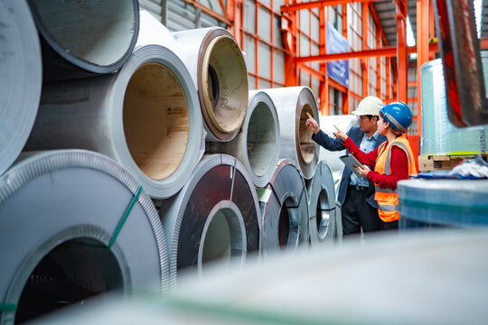 Factory manager and female engineer checking stacked steel coils together, discussing inventory, material quality, production planning and industrial warehouse operations.