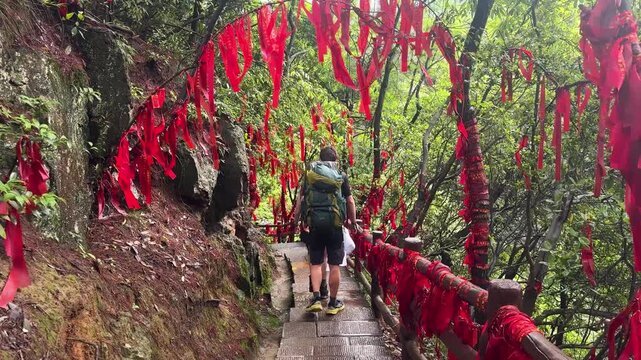 Tourists hiking on mountain path with red ribbons tied to trees. People walking along forest trail decorated with traditional prayer ribbons for luck.