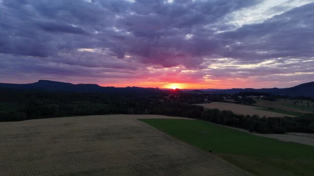 Fiery sunset glowing under heavy clouds above a dark forested valley and distant mesa