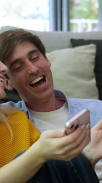 A happy young couple laughs hysterically while watching amusing content on a smartphone at home, sharing a joyful moment and connecting over digital entertainment.