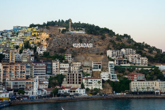 View of the Kusadasi sign on the hillside overlooking the coastal town with dense residential buildings, a clock tower, and the waterfront in Kusadasi, Aydin, Turkiye.