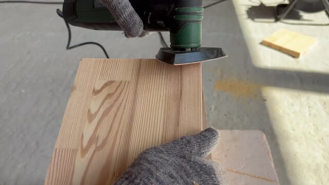 Close-up of a worker in gloves sanding the edge of a wooden board with a detail power sander in a workshop