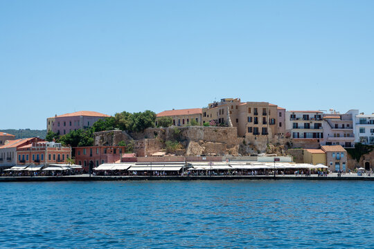 View of the historic Chania Old Town Marina with waterfront restaurants featuring white awnings and stone buildings under a clear blue sky in Chania, Crete, Greece.
