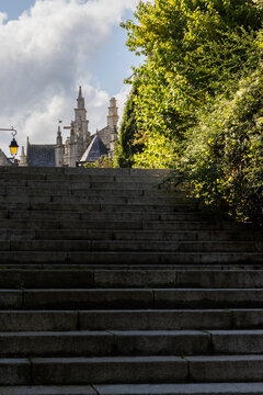 Wide stone steps leading up toward historic gothic architecture and lush green trees in the medieval town of Dinan, France, under a cloudy sky.