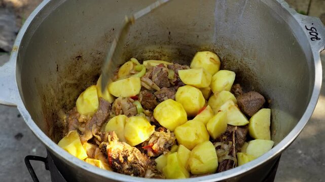 Traditional stew simmering in a large cast-iron kazan cauldron outdoors. Hot steam and rich ingredients highlight authentic ethnic cuisine