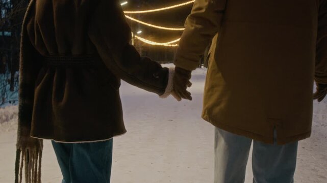 Young couple walking on snowy path, holding hands while camera tilts up from rear view. Footage ideal for winter holiday marketing and seasonal travel campaigns