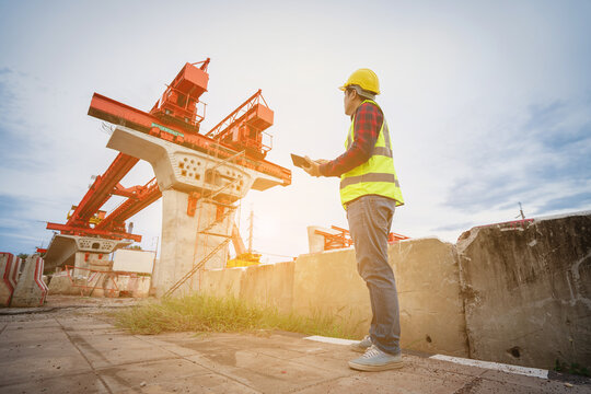 Asian civil engineers use tablets to record electronic data at a concrete highway bridge construction site. A male supervisor is inspecting things at the new concrete road and bridge construction site