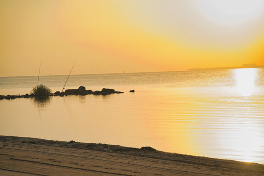 Golden sun dips low, casting warm glow. Ripples dance across calm, reflective water.  Beach sand stretches gently toward the sea